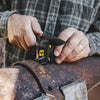 Person sharpening a folding knife blade using a compact black and yellow knife sharpener on a rusty metal surface, featured in Farm, Garden & Tool and Hand Tools and Hardware collections.