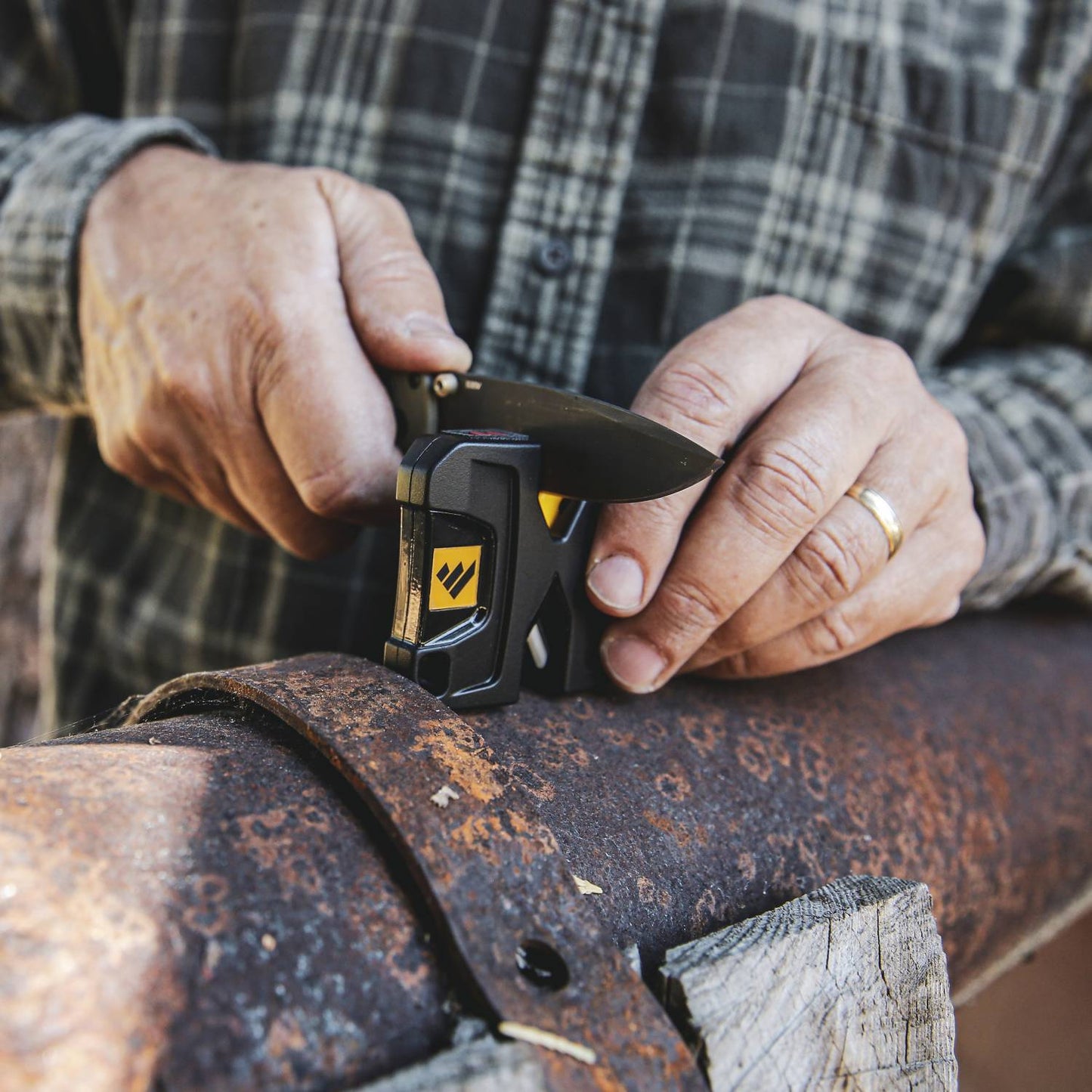 Person sharpening a folding knife blade using a compact black and yellow knife sharpener on a rusty metal surface, featured in Farm, Garden & Tool and Hand Tools and Hardware collections.