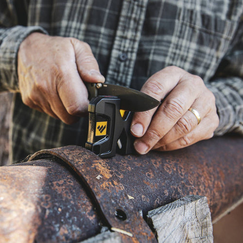 Person sharpening a folding knife blade using a compact black and yellow knife sharpener on a rusty metal surface, featured in Farm, Garden & Tool and Hand Tools and Hardware collections.