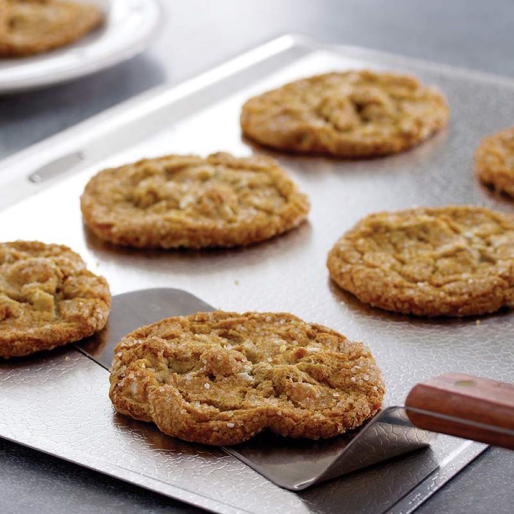 Freshly baked cookies cooling on a textured aluminum baking sheet with a wooden-handled spatula lifting one cookie, ideal for Kitchen & Food, Cookware, and Baking Supplies collections.