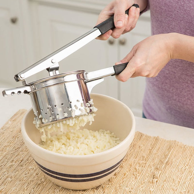 Hands using a stainless kitchen gadget to press cooked potatoes through holes into a ceramic bowl, demonstrating a classic cooking tool ideal for smooth mashed potatoes in a kitchen setting.