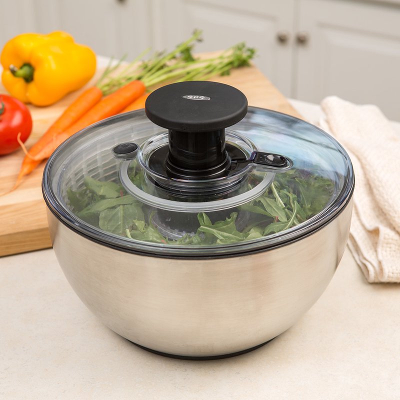 Brushed stainless steel salad spinner with clear locking lid and black push-button handle, containing fresh leafy greens, shown on kitchen countertop with fresh vegetables nearby, suitable for Kitchen & Food, Kitchen Gadgets, Cooking and Baking Helpers, Best Sellers, Promo Eligible collections.