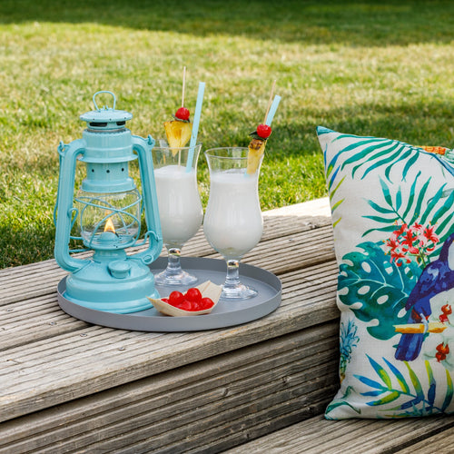 Handcrafted light blue galvanized oil lantern with thermal heat-resistant globe glowing softly on wooden decking next to tropical-patterned pillow, ideal for Lamps & Lighting, Camping, and Emergency Supplies collections.