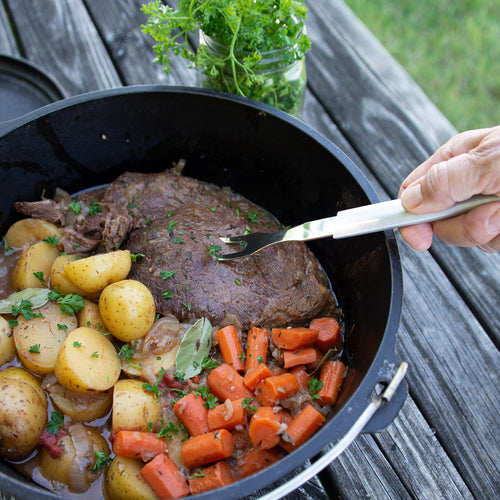Handheld carving fork with long stainless steel tines lifting cooked roast beef surrounded by seasoned potatoes and carrots in a cast iron skillet, ideal for Kitchen & Food, Storage and Serving, USA Made, Best Sellers, and Promo Eligible collections.