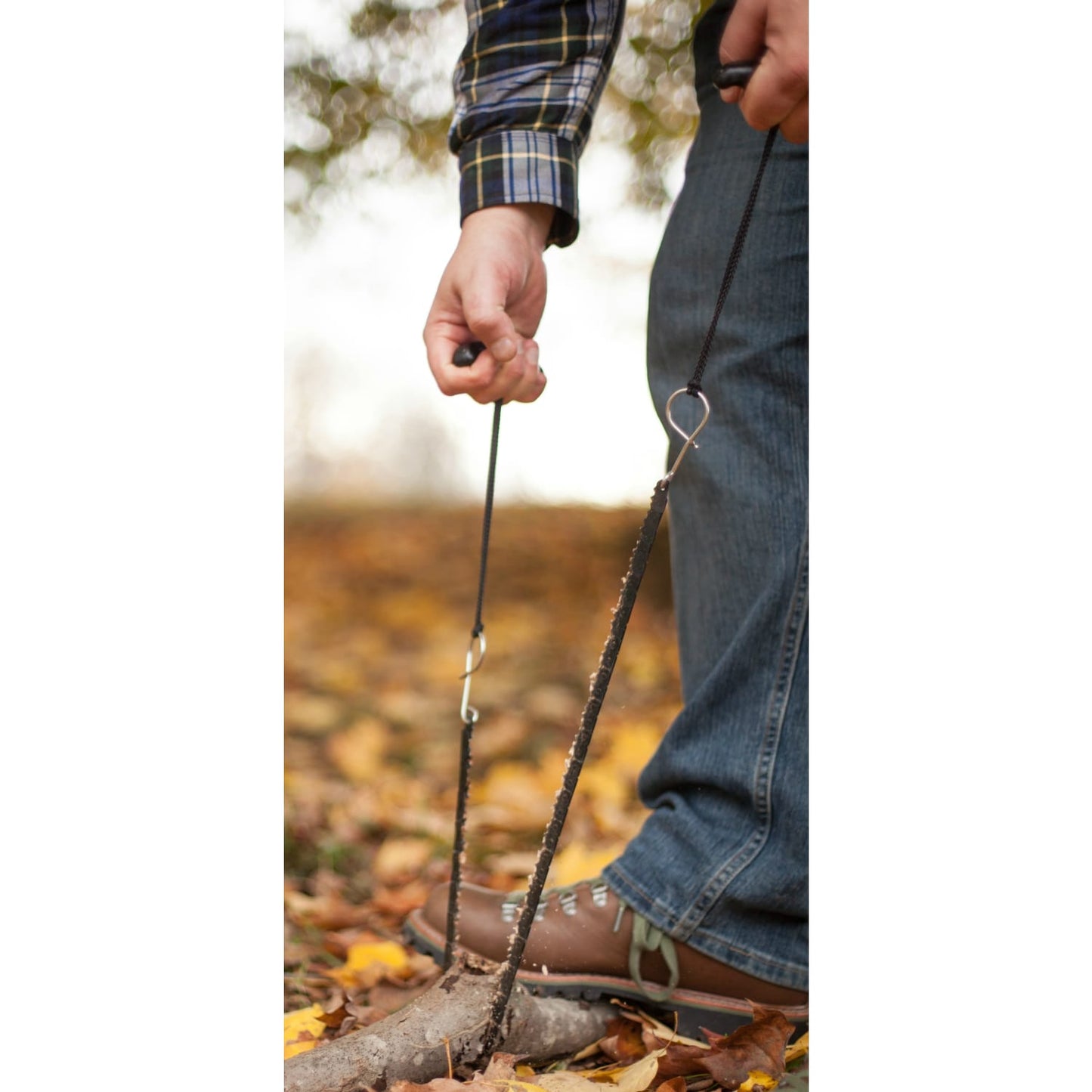 Person using a handcrafted 28-inch pocket chain saw with dual handles to cut a tree branch on a forest floor covered with autumn leaves, part of Farm, Garden & Tool and Wood Cutting collections.