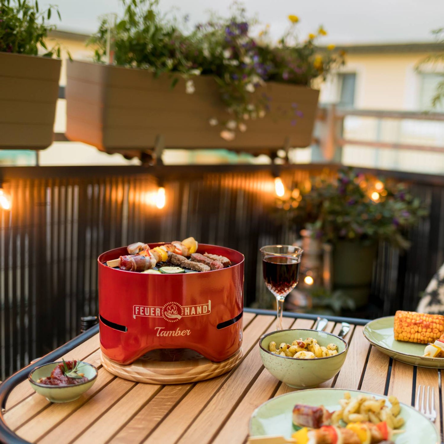 Unique handcrafted red tabletop charcoal grill cooking skewers on a wooden patio table, accompanied by side dishes and a glass of wine, ideal for Farm, Garden & Tool and Grilling collections.