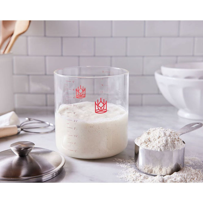 Clear borosilicate glass sourdough crock showing bubbly starter with red measurement markings next to a metal measuring cup filled with flour and stainless steel lid nearby, suited for Kitchen & Food, Cookware, Baking Supplies, Storage and Serving, and Best Sellers collections.