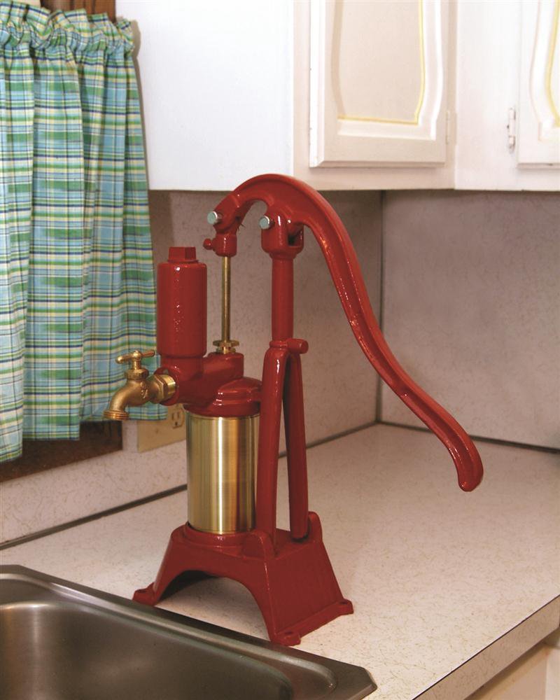 Red pressurized water pump with brass cylinder and spout, designed for steady water flow, displayed on a kitchen counter beside a sink and plaid curtains in a USA made appliances collection.