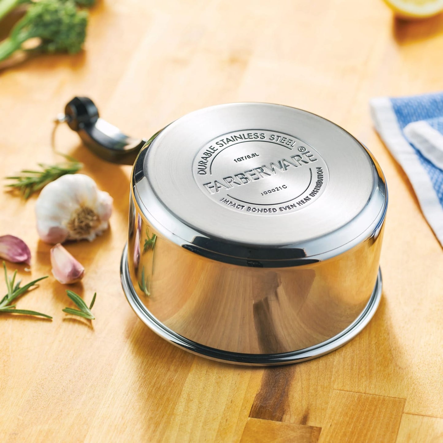 Shiny stainless steel saucepan with heavy-duty base embossed with branding, resting upside down on a wooden kitchen counter surrounded by garlic and fresh herbs, part of cookware collections.