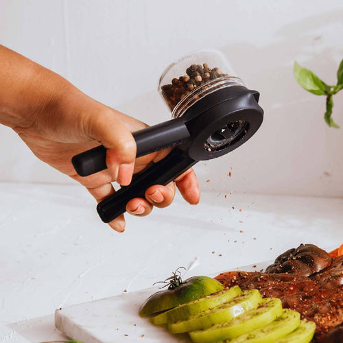 Hand holding black Ortwo Lite Salt and Pepper Mill grinding whole peppercorns over sliced green tomatoes and seasoned meat, featured in Kitchen & Food and Kitchen Gadgets collections.