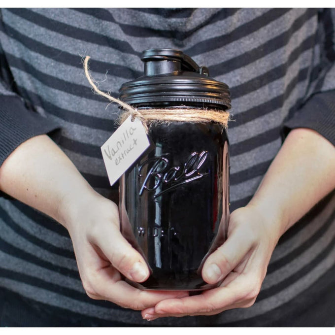 Person holding a wide-mouth glass jar filled with dark liquid, sealed with a black pour lid featuring a carry loop, tagged with "Vanilla extract," in a cozy striped sweater, suitable for Kitchen & Food, Dinnerware, Kitchen Gadgets, Kitchen Accessories, USA Made, Best Sellers, and Promo Eligible collections.