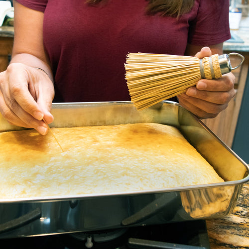 Handcrafted miniature broom cake tester with a metal loop for hanging, shown held by hand while checking doneness of a freshly baked cake in a stainless steel pan, part of Kitchen & Food, Cleaning Utensils and Gadgets collections made in USA.