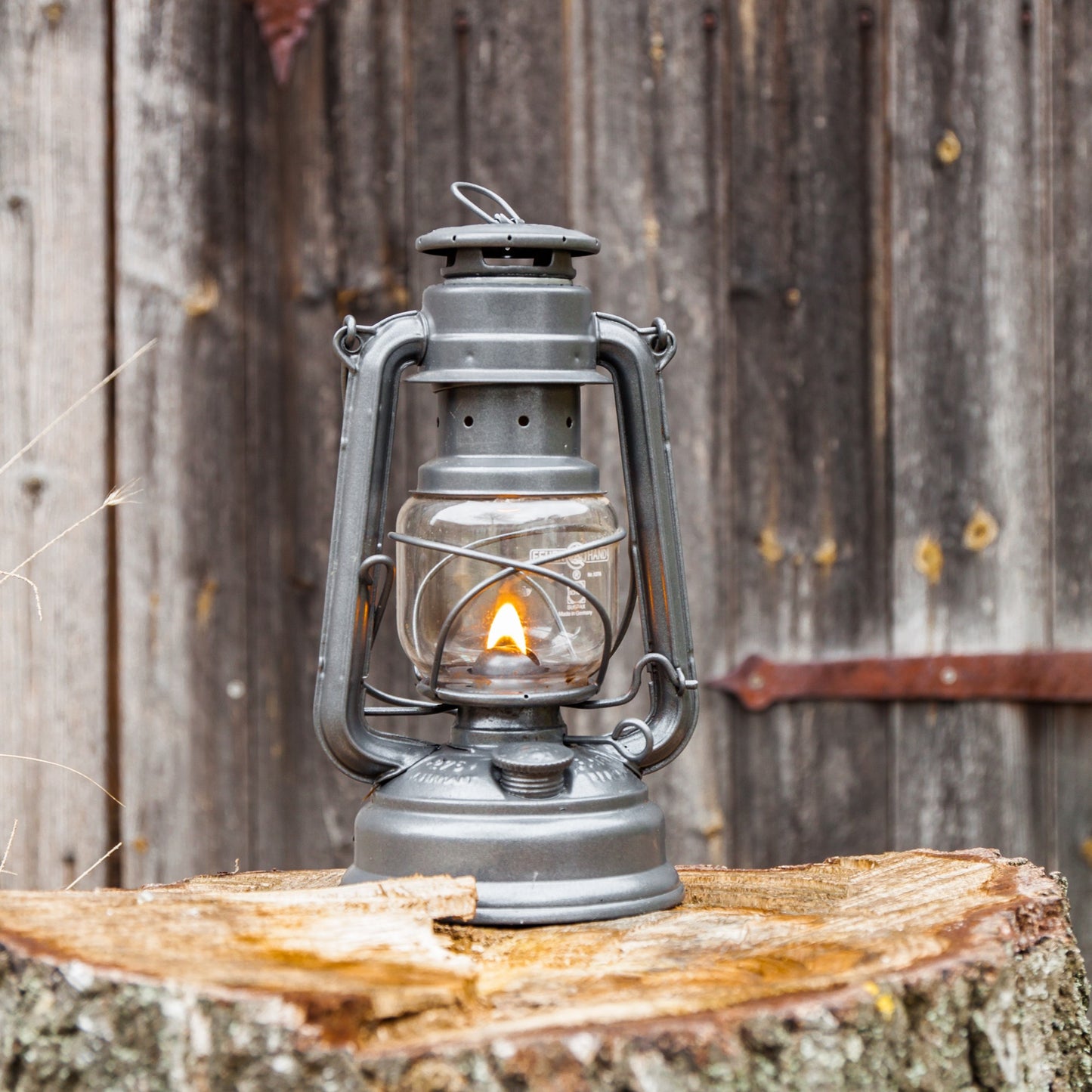 Handcrafted galvanized oil lantern with durable thermal heat-resistant globe softly glowing on a rustic tree stump against weathered wooden background, ideal for Lamps & Lighting, Camping, and Emergency Supplies collections.