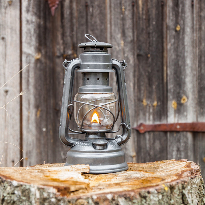Handcrafted galvanized oil lantern with durable thermal heat-resistant globe softly glowing on a rustic tree stump against weathered wooden background, ideal for Lamps & Lighting, Camping, and Emergency Supplies collections.