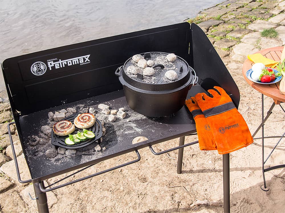 Outdoor cooking setup featuring a sturdy steel table with a cast iron Dutch oven and grill grate topped with hot coals, paired with orange protective gloves, ideal for camping and garden use.