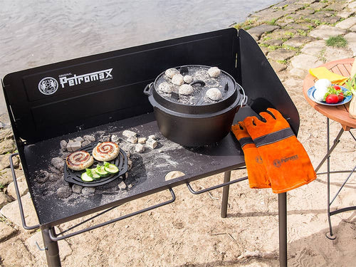 Outdoor cooking setup featuring a sturdy steel table with a cast iron Dutch oven and grill grate topped with hot coals, paired with orange protective gloves, ideal for camping and garden use.