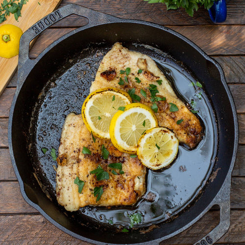 Handcrafted cast iron pan with dual handles, cooking seasoned fish fillets garnished with lemon slices and fresh herbs, featured in Camping, Kitchen & Food, and USA Made collections.