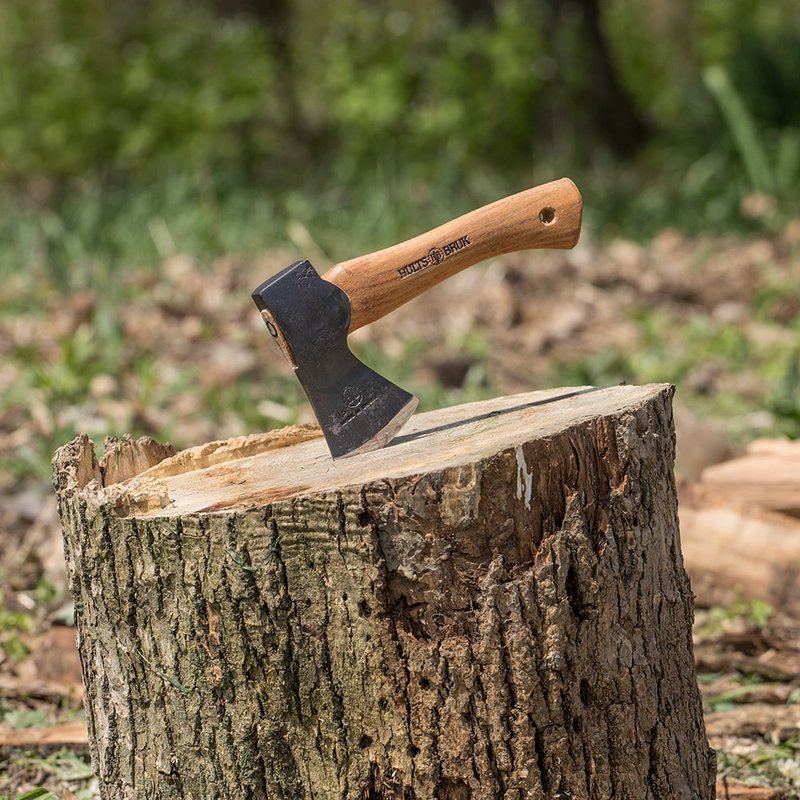 Hand-forged Swedish steel hatchet with a curved wooden handle embedded in a tree stump, crafted for camping, farm, and woodcutting tasks, featuring a protective leather sheath option.