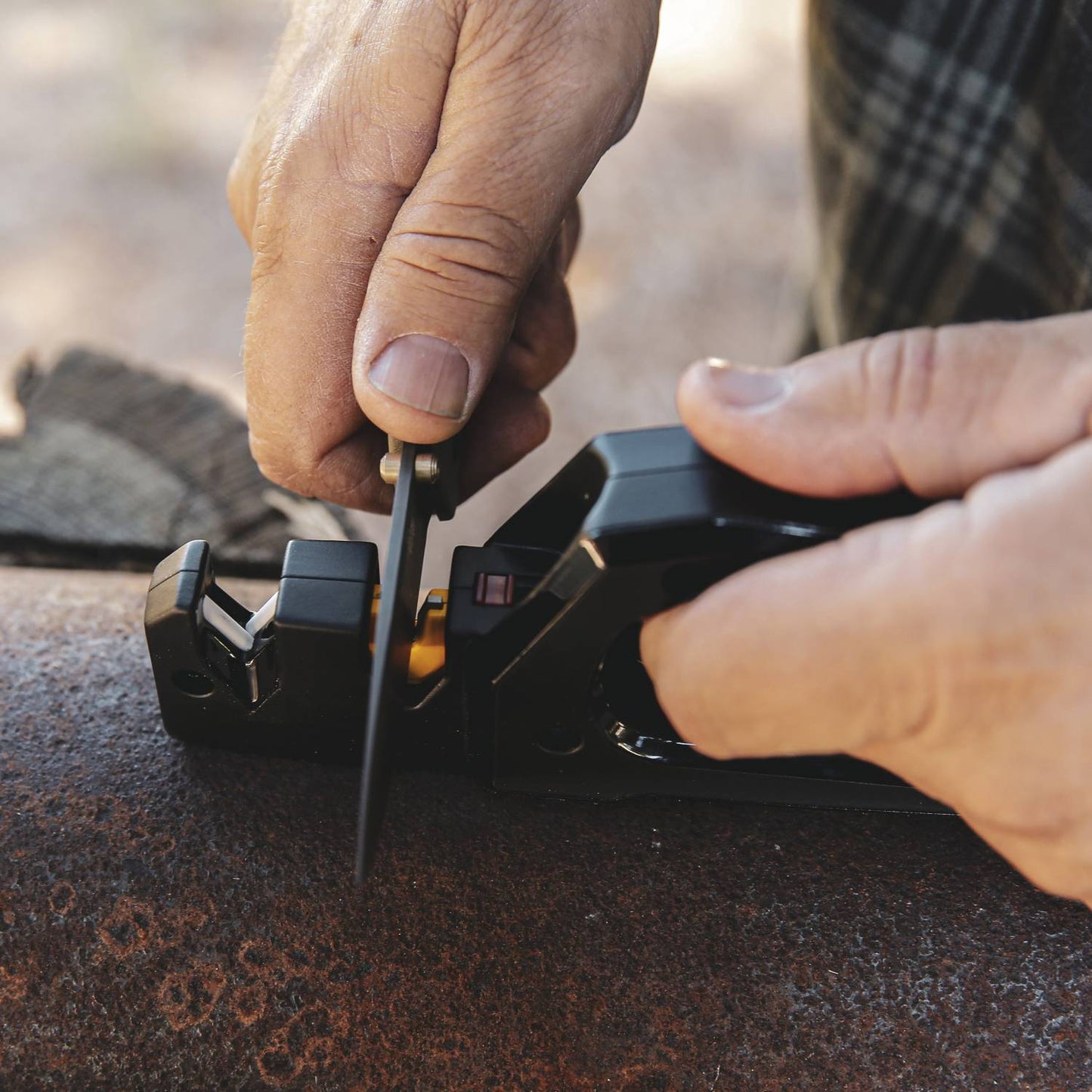 Person using a black handheld sharpener to quickly restore a knife blade outdoors on a rustic wood surface, suited for Farm, Garden & Tool, Hand Tools and Hardware, and Sharpening collections.