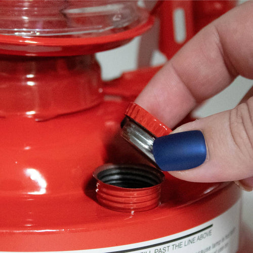 Close-up of a hand with matte blue nail polish unscrewing the red metal fuel cap on a handcrafted red oil lantern, suitable for camping, pest control, and outdoor lighting in farm and garden settings.