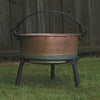 Copper kettle resting on a sturdy three-legged iron stand with a curved handle, designed for stable use in farm, camping, and kitchen settings, made in USA.