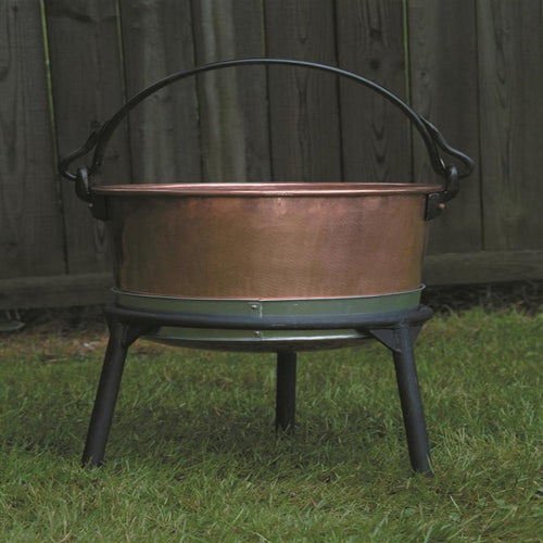 Copper kettle resting on a sturdy three-legged iron stand with a curved handle, designed for stable use in farm, camping, and kitchen settings, made in USA.