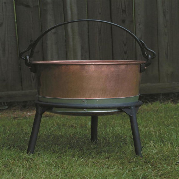 Copper kettle resting on a sturdy three-legged iron stand with a curved handle, designed for stable use in farm, camping, and kitchen settings, made in USA.