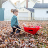Young child pushing a handcrafted red Eli & Mattie kids wheelbarrow with hardwood handles through a yard covered in autumn leaves, promoting outdoor play and garden fun.