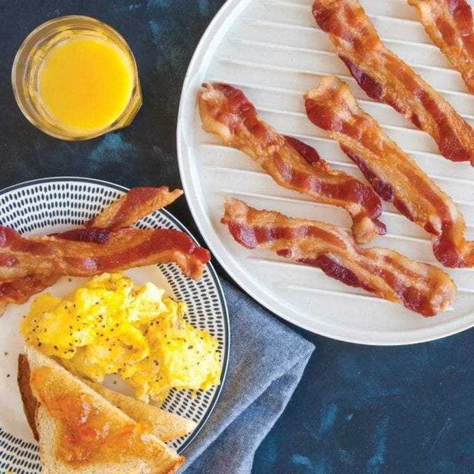 Crisp cooked bacon strips resting on a ribbed white plate designed to drain grease, accompanied by scrambled eggs, toast with jam, and a glass of orange juice, highlighting a handcrafted kitchen gadget from USA Made collections.