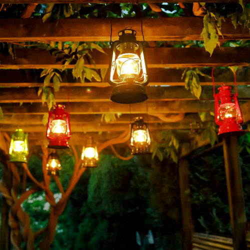 Handcrafted galvanized oil lanterns glowing warmly in various colors, hanging from a rustic wooden pergola surrounded by greenery, ideal for Lamps & Lighting, Camping, and Emergency Supplies collections.