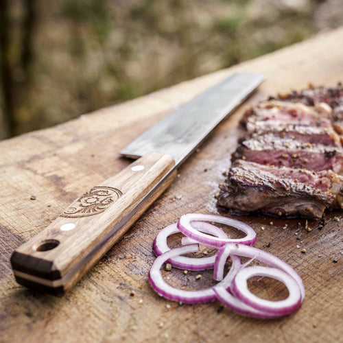 Handcrafted outdoor chef’s knife with a sturdy walnut handle and sharp steel blade resting on a wooden board next to sliced grilled meat and fresh red onion rings, ideal for Farm, Garden & Tool and Camping collections.