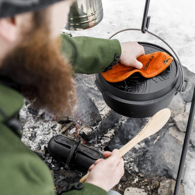 Bearded person wearing green jacket lifting a cast iron Dutch oven from an outdoor campfire using an orange heat-resistant glove, with a wooden spoon and firepit stones nearby, suited for Farm, Garden & Tool and Camping collections.