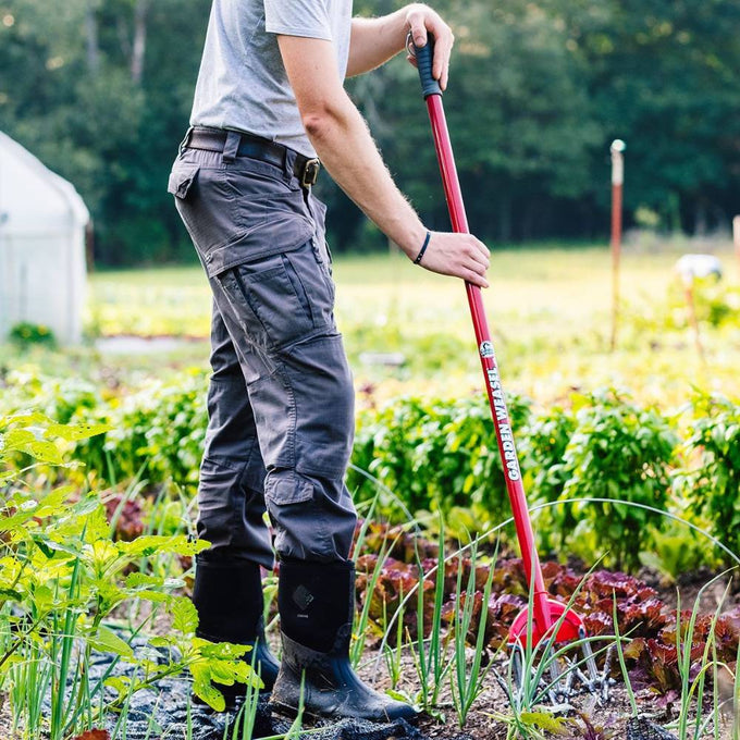 Person using a long-handled garden cultivator with rust-resistant serrated tines and a comfort-grip handle to loosen soil and remove weeds in a lush vegetable garden, featured in Farm, Garden & Tool and Digging and Cultivating collections.