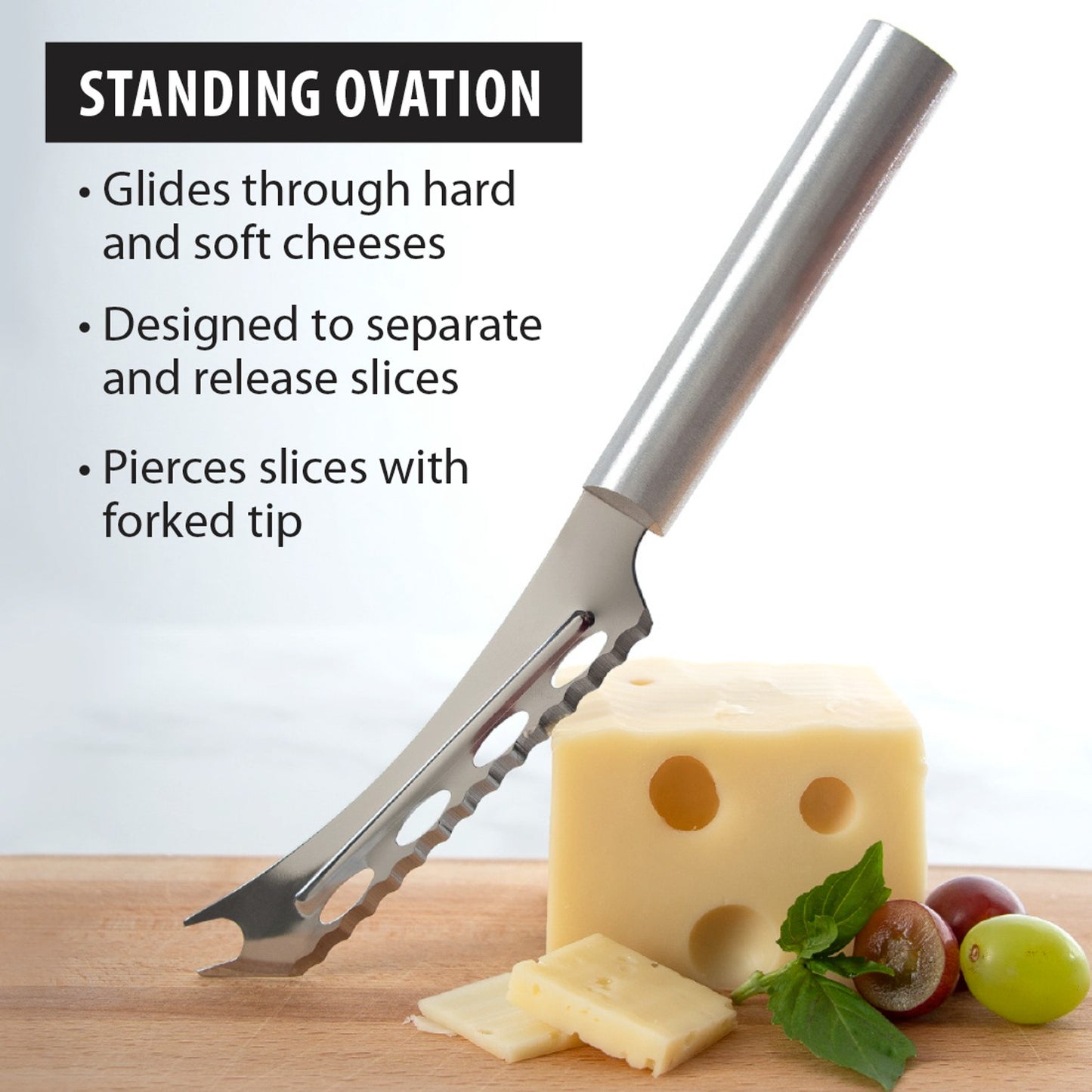 Serrated cheese knife with stainless steel blade and forked tip slicing through Swiss cheese with grapes and herbs nearby, a handcrafted USA made kitchen tool ideal for cheese and food prep.