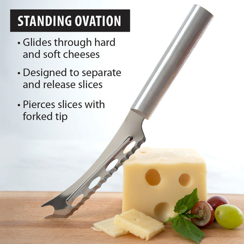 Serrated cheese knife with stainless steel blade and forked tip slicing through Swiss cheese with grapes and herbs nearby, a handcrafted USA made kitchen tool ideal for cheese and food prep.