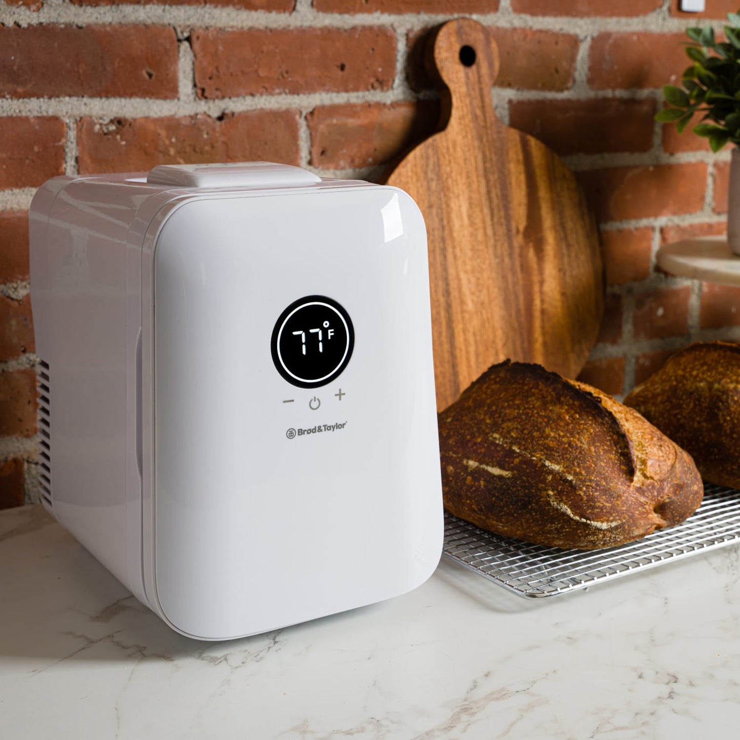 Compact white temperature-controlled device displaying 77°F on a digital screen, placed on a marble countertop next to freshly baked artisan bread and a wooden cutting board, featured in Kitchen & Food and Baking Supplies collections.
