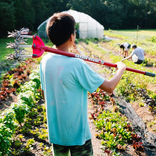 Person holding a long-handled garden tool with rust-proof serrated tines and comfort-grip handle, working in a vegetable garden surrounded by growers, featured in Farm, Garden & Tool collections.