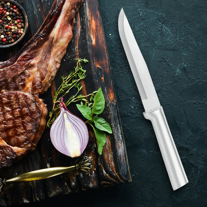 Handcrafted Rada Stubby butcher knife with a 5-3/8" stainless steel blade and silver brushed aluminum handle beside grilled meat, fresh herbs, and a halved red onion on a dark textured surface, ideal for Farm, Garden & Tool and Kitchen & Food collections.