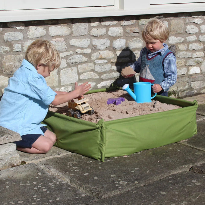 Two young children playing with toys in a green raised fabric sandpit filled with sand outdoors, suitable for outdoor play, children's toys, garden accessories, and promo collections.