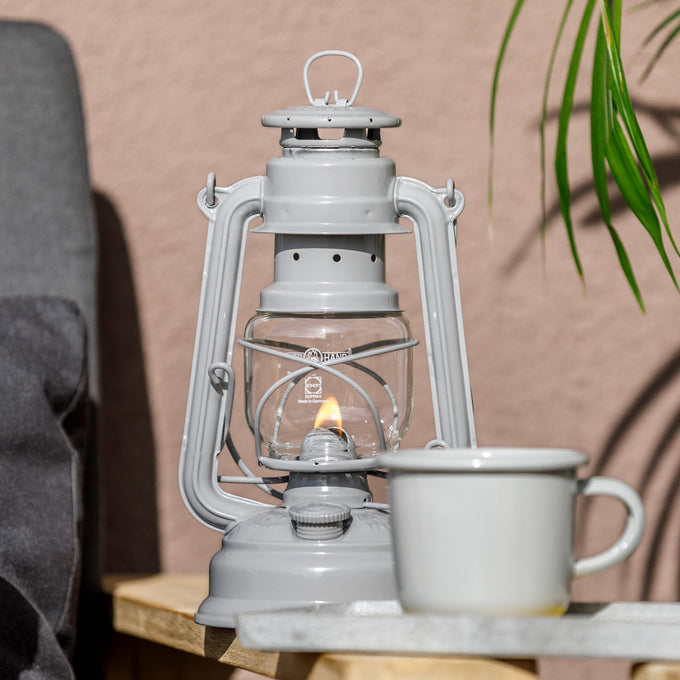 Handcrafted pale gray galvanized oil lantern with thermal heat-resistant globe, softly lit next to a ceramic mug on a wooden surface, ideal for Lamps & Lighting, Camping, and Emergency Supplies collections.