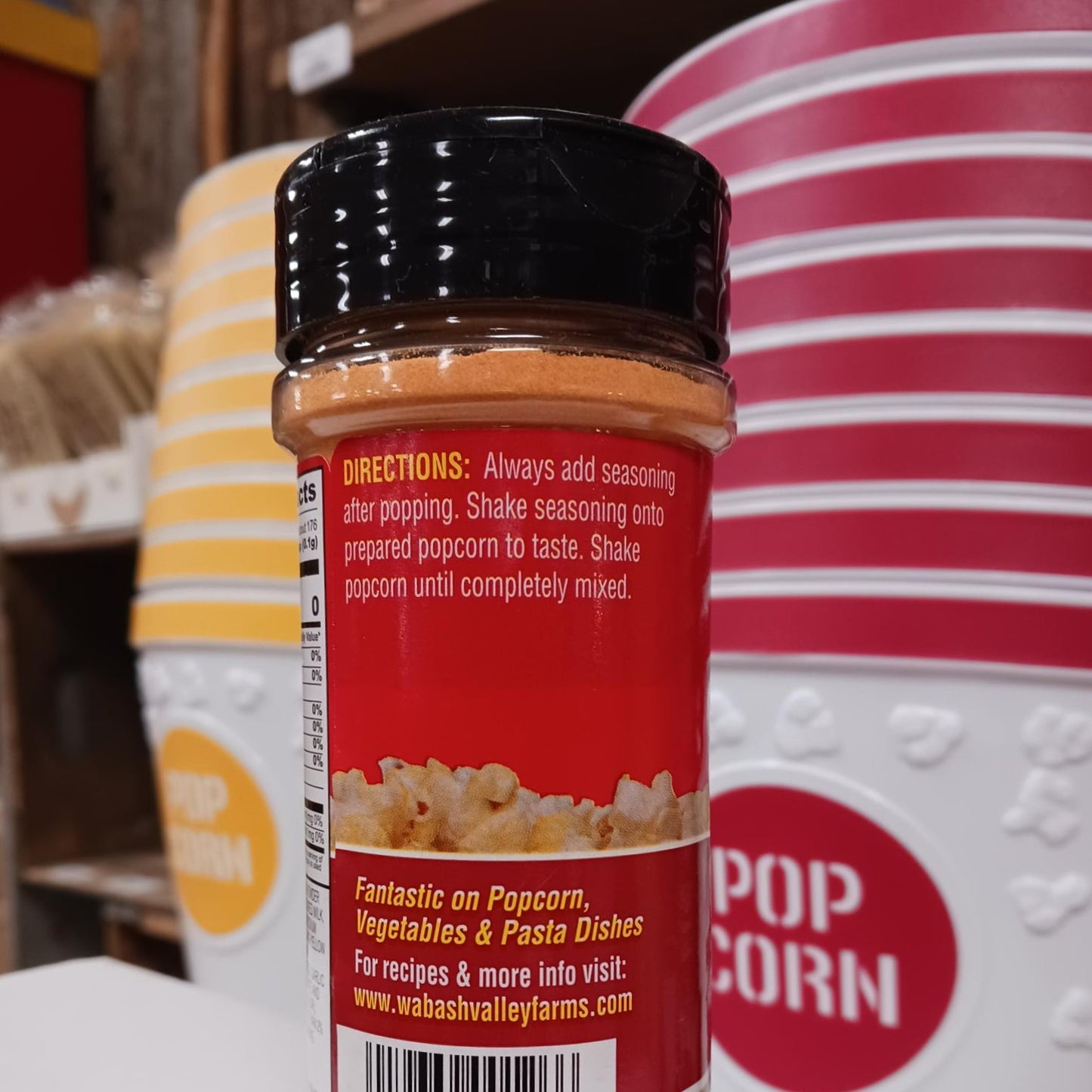 Clear plastic bottle of orange cheesy popcorn seasoning with black lid showing usage directions, set against a background of striped popcorn containers in a kitchen display.