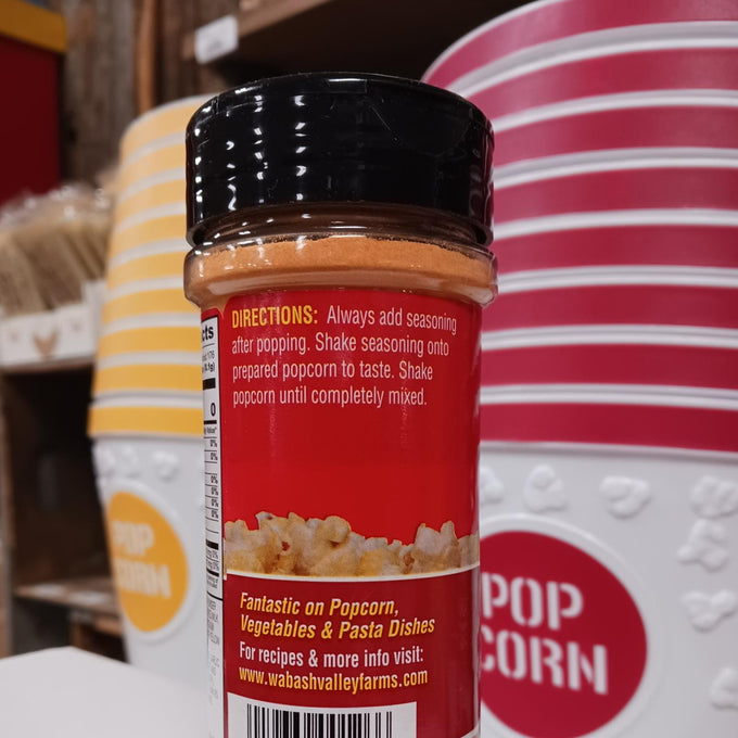 Clear plastic bottle of orange cheesy popcorn seasoning with black lid showing usage directions, set against a background of striped popcorn containers in a kitchen display.