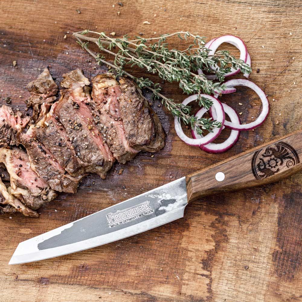 Handcrafted utility knife with a hardened steel blade and engraved walnut handle next to sliced steak, red onion rings, and fresh herbs, ideal for kitchen, camping, and food prep.