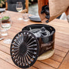 Black charcoal pieces being poured from a paper bag into a compact, black tabletop grill with a decorative vented lid resting on a wooden table, suitable for grilling and outdoor cooking collections.