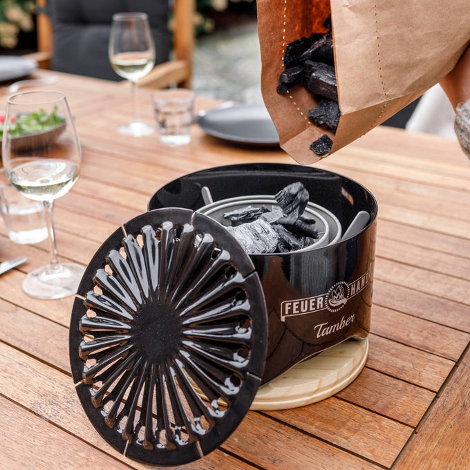 Black charcoal pieces being poured from a paper bag into a compact, black tabletop grill with a decorative vented lid resting on a wooden table, suitable for grilling and outdoor cooking collections.