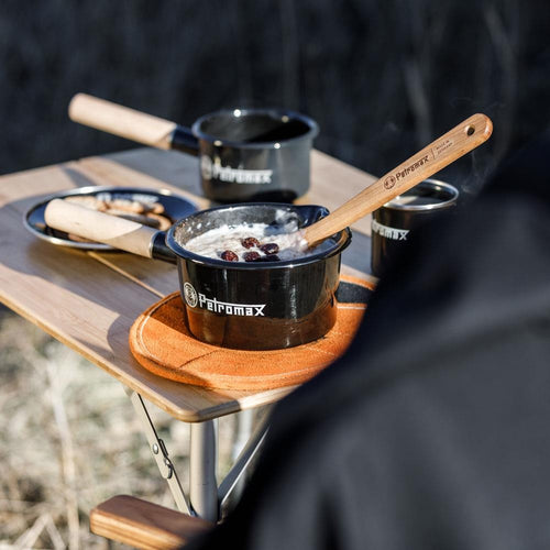 Black enamel pan with wooden handle and petromax logo cooking oatmeal with raisins, placed on a wooden outdoor table alongside matching cookware, suited for camping and outdoor use in Farm, Garden & Tool, Kitchen & Food, and Camping collections.