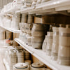 Shelves filled with handcrafted stoneware mugs and kitchenware in natural tones with subtle blue stripes, featured in USA Made, Kitchen & Food, and Dinnerware collections.
