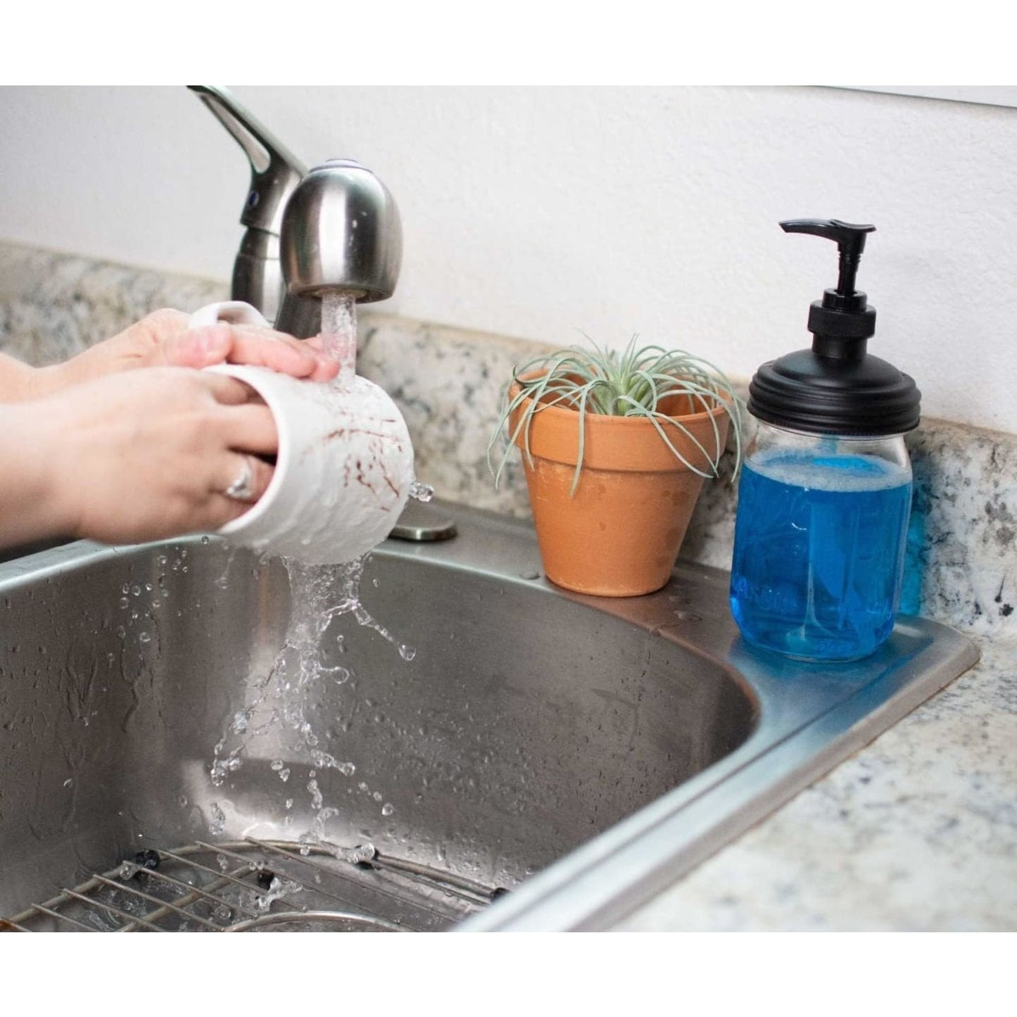 Hand using water to rinse a white ceramic mug over a stainless steel sink next to a regular-mouth mason jar soap pump filled with blue liquid soap, set on a granite countertop with a small potted plant nearby, ideal for Health & Wellness and Home & Decor collections.