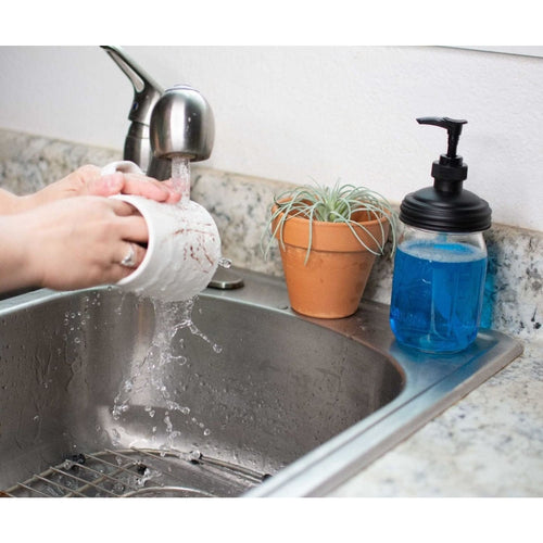 Hand using water to rinse a white ceramic mug over a stainless steel sink next to a regular-mouth mason jar soap pump filled with blue liquid soap, set on a granite countertop with a small potted plant nearby, ideal for Health & Wellness and Home & Decor collections.