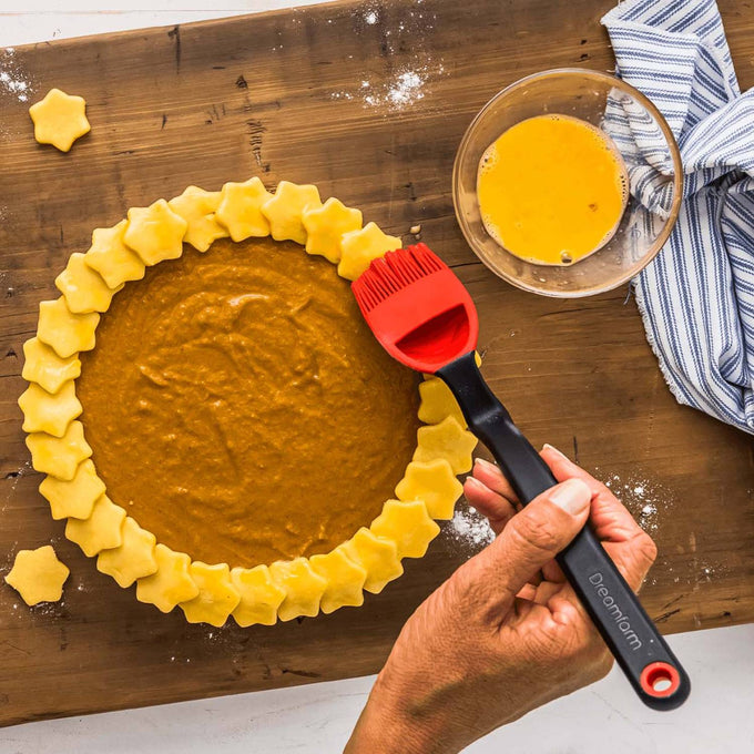 Hand holding a red and black silicone basting brush with ergonomic handle, applying egg wash to star-shaped pie crust edges on a wooden surface with bowl and striped towel, perfect for Kitchen & Food and Cooking and Baking Helpers collections.